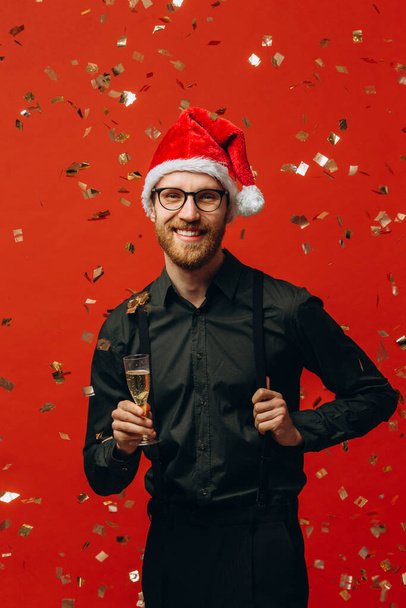 Χριστούγεννα, πάρτι και γιορτές. Όμορφος άντρας με καπέλο Σάντα υψώνει ένα ποτήρι σαμπάνια και χαμογελά, γιορτάζει την Πρωτοχρονιά, κόκκινο φόντο. - Φωτογραφία, εικόνα