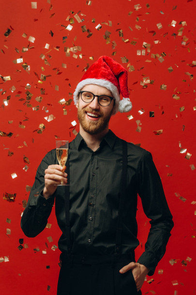 Noël, fête et concept de vacances. Charmant homme au chapeau de Père Noël levant un verre de champagne et souriant, célébrant le Nouvel An, fond rouge. - Photo, image