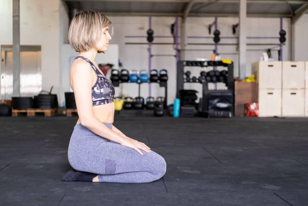 Une femme blonde mi-adulte méditant à genoux ou vajrasana pose les yeux fermés pendant sa pratique de yoga en flux vinyasa seule et portant des leggings et un soutien-gorge de sport dans une salle de gym. - Photo, image