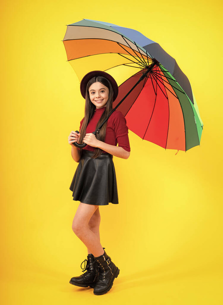 Fashion autumn weather for children. Teenager girl with umbrella in autumn skirt, sweater, boots and hat isolated on yellow studio background. Autumn trendy teen kids clothes - Φωτογραφία, εικόνα