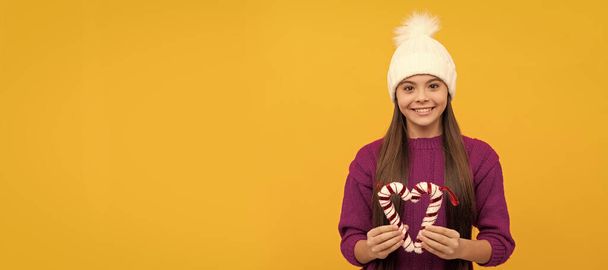 Ευτυχισμένο το νέο έτος. Ευτυχισμένο παιδί με χειμερινό καπέλο. Παιδί με ζαχαρωτά. Έφηβη με κίτρινο φόντο. Οριζόντια αφίσα του απομονωμένου προσώπου του παιδιού, κεφαλίδα banner, αντίγραφο χώρου - Φωτογραφία, εικόνα