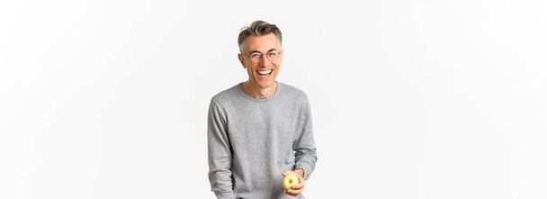 Obrázek šťastného a zdravého muže středního věku, jak drží zelené jablko, směje se a bezstarostně se usmívá, stojí nad bílým pozadím. - Fotografie, Obrázek