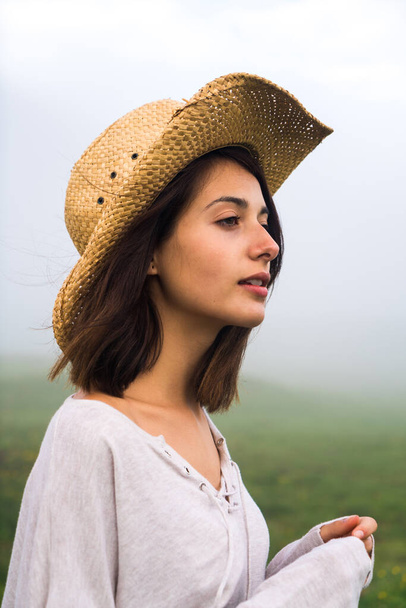 Portrait of a caucasian woman wearing a straw hat looking away from camera on a foggy background. - Фото, изображение