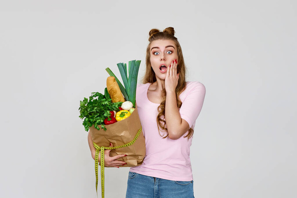 surprised caucasian vegetarian woman in pink t-shirt holds paper bag with groceries after shopping on gray background. copy space - Foto, immagini