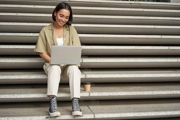 Портрет молодої азіатки, яка використовує свій ноутбук, сидить на відкритому повітрі на сходах. Щаслива усміхнена дівчина з персональним комп'ютером
. - Фото, зображення
