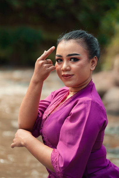 Krásná Javanese žena s make-upem a fialové šaty pózovat s rukou, zatímco sedí na skále u řeky uvnitř vesnice - Fotografie, Obrázek