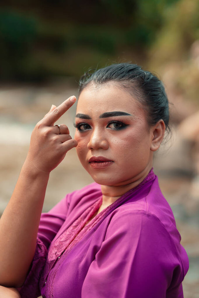 Bella donna giavanese con trucco e vestito viola in posa con la mano mentre sedeva sulla roccia vicino al fiume all'interno del villaggio - Foto, immagini
