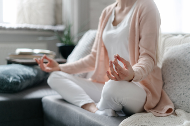 Achtsamkeitsmeditation und Konzept für psychische Gesundheit. Junge Frau in lässiger Kleidung praktiziert Yoga zu Hause, sitzt in Lotus-Pose auf der Couch im Wohnzimmer und ruht sich nach einem anstrengenden Arbeitstag aus - Foto, Bild