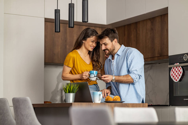 キッチンでコーヒーを飲みながら仕事でノートパソコンを使うカップル. - 写真・画像