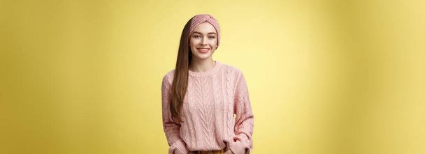 Charming friendly young girl in knitted sweater and headband holding hands in pockets, smiling, looking positive motivated achieving goals having great day, posing positive against yellow background. - Photo, Image