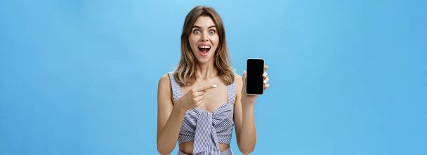 Aufgeregt kaufte die glückliche Frau mit klappernden Zähnen schließlich ein nagelneues Smartphone-Haltegerät in der Hand und zeigte auf den Handybildschirm, auf dem eine coole App vor Freude vor blauer Wand breit lächelte. Werbung - Foto, Bild
