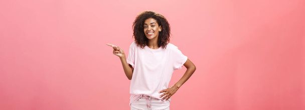 Hé, kijk. Portret van schattige chill en vriendelijke stijlvolle Afrikaanse Amerikaanse vrouw met afro kapsel hand in hand op taille wijzend naar rechts en glimlachend vrolijk over roze achtergrond. Kopieerruimte - Foto, afbeelding
