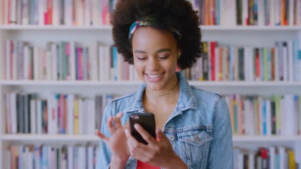 Telefon, közösségi média és egy fekete diáklány egy egyetemi egyetemi könyvtárban. Mobil, kommunikáció és tanulás egy női tanulóval egy könyvesboltban tanulásra. - Felvétel, videó
