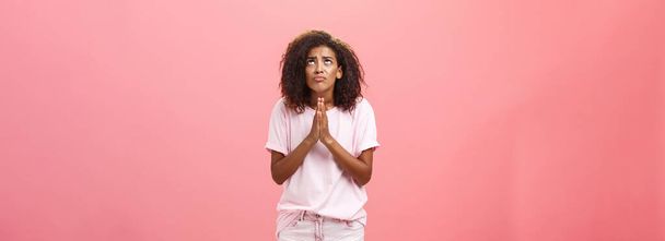 Studioaufnahme unglücklicher elender und hoffnungsloser süßer afrikanisch-amerikanischer Frau, die die Hände im Gebet nahe der Brust hält und mit ernst dreinblickender besorgter Miene aufblickt und sich Gott über rosa Wand wünscht - Foto, Bild