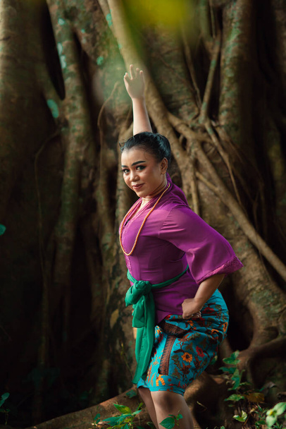 Asian woman takes a dancing pose in the jungle while wearing a purple dress and a golden necklace with a beautiful smile on her face - Photo, Image