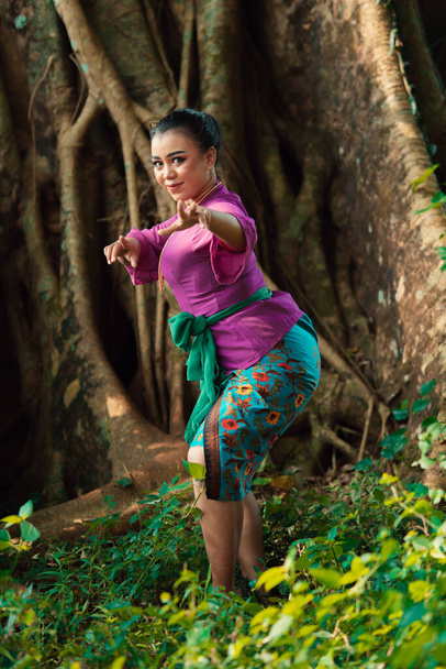 Sexy Asian woman dancing pose while wearing a beautiful purple dress and green belt on her body in front of the big brown tree inside the forest - Fotografie, Obrázek