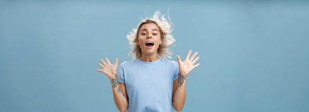 Creative happy and playful beautiful blond girl with tattoos on arms raising palms up opening mouth facing wind while hair strands flicking on air jumping having fun over blue background. Emotions - Photo, Image