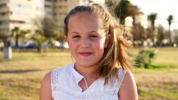 Kind, glimlach en vrolijk gezicht op een stadspark met wind in het haar, geluk en vrijheid om buiten te spelen op gras met bomen en plezier. Portret van een meisje op vakantie in Zuid-Afrika met tanden lachen. - Video