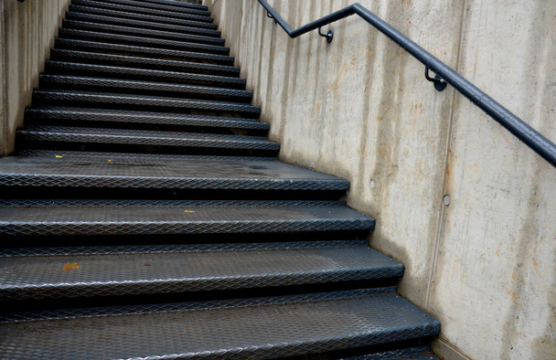 betonowe ściany, pomiędzy którymi zaklinowane są metalowe schody o wzorze antypoślizgowym. poręcz na odsłoniętym betonie jest pomalowana na czarno. - Zdjęcie, obraz