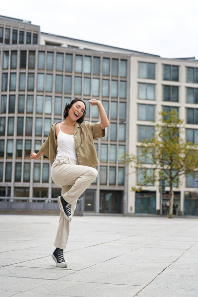 Vertical shot of dancing girl in headphones, listening music on street, enjoying sound in earphones. - Zdjęcie, obraz