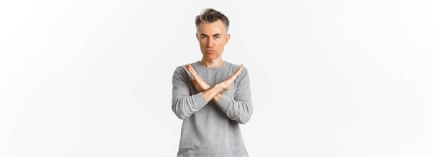 Image of angry middle-aged man looking serious, making cross gesture to stop something bad, telling no, prohibit action, standing over white background. - Photo, Image