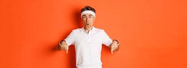 Concept of workout, sports and lifestyle. Portrait of surprised middle-aged male athlete, wearing clothes for gym, pointing fingers down and looking amazed, standing over orange background. - Photo, Image