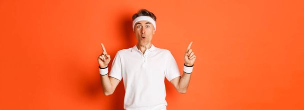 Porträt eines gutaussehenden Mannes mittleren Alters in Sportkleidung, der mit erhobenem Zeigefinger ein Werbebanner über Fitnessstudio oder Workout zeigt, vor orangefarbenem Hintergrund stehend. - Foto, Bild