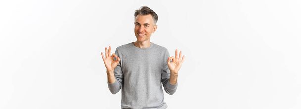 Portrait of handsome middle-aged man, smiling and looking confident while showing okay signs, guarantee something is good, standing over white background. - Photo, Image