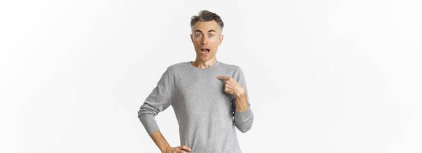 Portrait of handsome middle-aged gay man looking surprised, pointing at himself, being chosen, standing over white background in grey basic sweater. - Zdjęcie, obraz