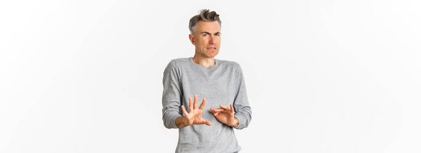 Portrait of reluctant middle-aged man avoiding something bad, shaking hands and declining offer, grimacing from disgust, standing over white background. - Photo, Image