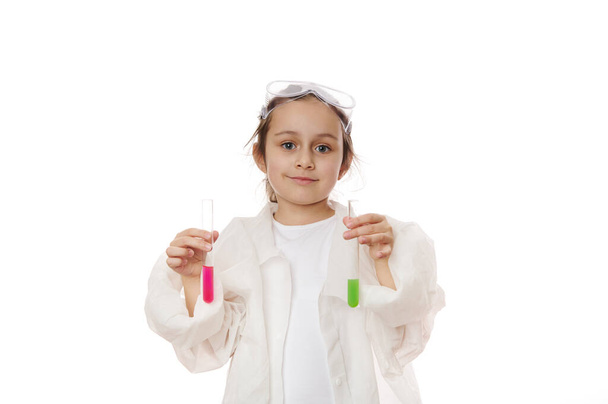 Lovely child girl, future chemist scientist in lab coat, smiles looking at camera, holding out test tubes with liquid chemical, isolated on white background. Kids learning chemistry. Education concept - Foto, afbeelding