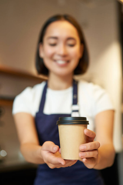 Lächelnde asiatische Barista, die dem Kunden Befehl gibt, Kaffeetasse zum Mitnehmen hält, Schürze trägt, hinter dem Tresen im Café arbeitet. - Foto, Bild