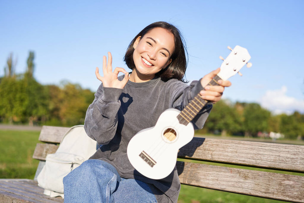 Cute smiling girl shows ok sign and her new ukulele, sits on bench in park, recommends musical instrument. - Fotografie, Obrázek