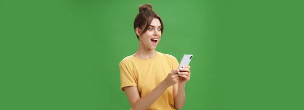 Kobieta czyta zaskakujące satysfakcjonujące wiadomości w smartfonie otwarcia ust z podniecenia, uśmiechając się zdumiony patrząc zdumiony na ekranie telefonu komórkowego pozowanie na zielonym tle w casual żółty t-shirt - Zdjęcie, obraz
