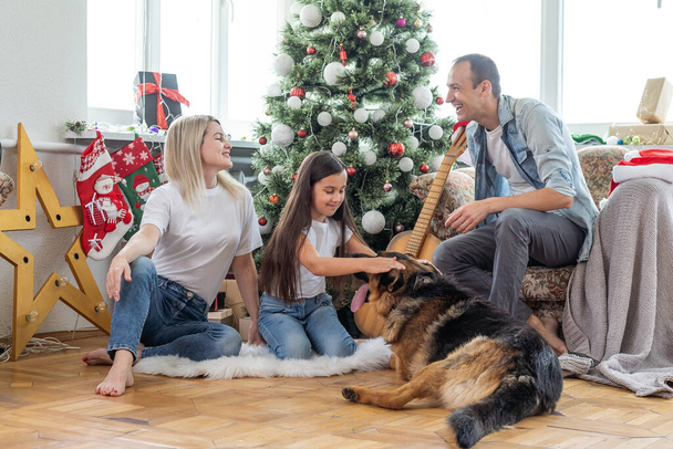 χαμογελαστή οικογένεια και κόρη με το σκυλί κάθεται κοντά στο χριστουγεννιάτικο δέντρο με δώρα. - Φωτογραφία, εικόνα