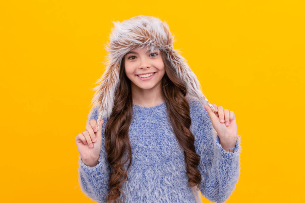 冬のファッション。耳フラップ付きの帽子を被った幸せな子供。女性ファッションモデル。黄色の背景にニットを着た10代の女の子。暖かい服を着た子供の肖像画です。肯定的な感情を. - 写真・画像