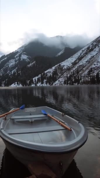 鏡水で森の中の山の湖。白いボートと木製の桟橋。水は曇りの空、雪の山や峰、針葉樹林の会社の風景を反映します。カザフスタンのコルサイ湖 - 映像、動画