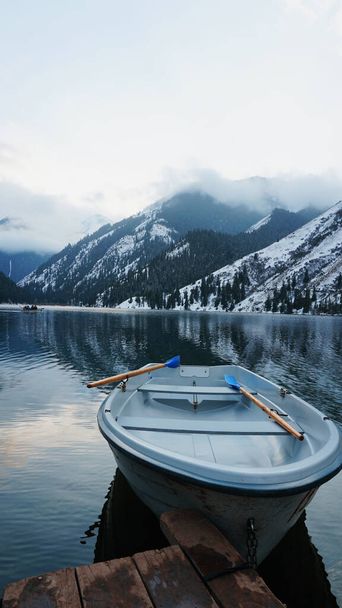 Un lac de montagne dans la forêt avec de l'eau en miroir. Masse en bois avec bateaux blancs. L'eau reflète le paysage d'un ciel nuageux, de montagnes enneigées et de sommets, de sapins résineux. Lac Kolsai, Kazakhstan - Photo, image