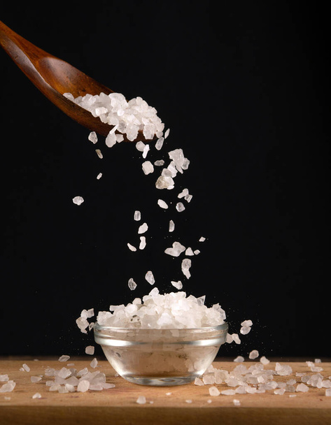 Large white salt is poured from a wooden spoon into a glass salt cellar. Copy space on a black background - Photo, Image
