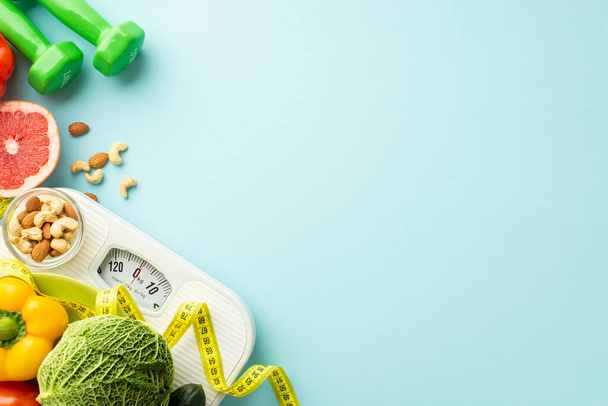 Weight loss concept. Top view photo of scales dumbbells vegetables fruits almonds cashew and tape measure on isolated pastel blue background with empty space - Photo, Image