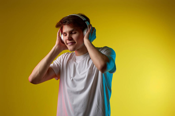 Young man listening to music with wireless headphones on yellow background. Neon lighting - Fotografie, Obrázek