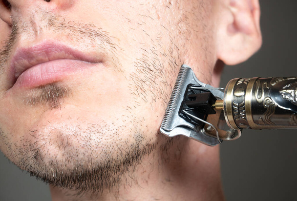 Mies leikkaa parran Electric metalli partaveitsi tai kuiva parranajokone. Leikkuri leikkaamiseen. Lyhyt, harva parta ihmisten kasvoilla. Hiusten kasvuongelmia. Parran karvaiset harjakset. - Valokuva, kuva