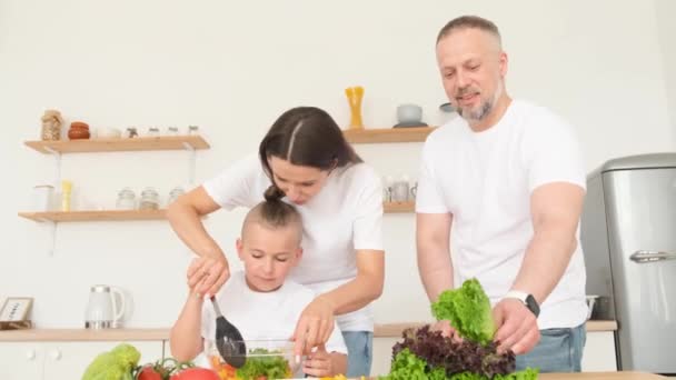 Pai, a mãe e o filho estão a preparar uma salada saudável de vegetais frescos juntos. Família em uma cozinha branca moderna. Casa de família olhar - Filmagem, Vídeo