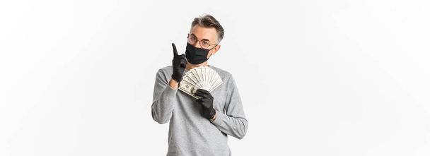 Concept van covid-19, sociale afstand en levensstijl. Portret van een gelukkige man van middelbare leeftijd in medisch masker, handschoenen en bril, met geld en wijzende vinger omhoog, glimlachend blij. - Foto, afbeelding