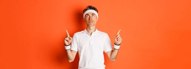 Portrait of handsome middle-aged male athlete, wearing sports clothing, looking and pointing fingers up at promo banner, standing against orange background. - Foto, Imagem