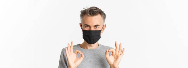 Concept of covid-19, social distancing and quarantine. Close-up of confident middle-aged man, wearing black medical mask, assure everything good, showing okay signs, white background. - Valokuva, kuva