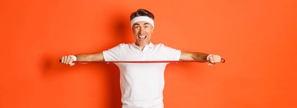 Concept d'entraînement, gymnase et mode de vie. Homme de condition physique actif et en bonne santé d'âge moyen, tenant la corde à étirer pour les exercices et souriant, debout sur fond orange. - Photo, image