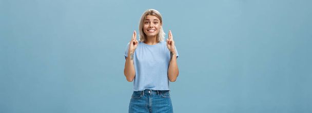 Hope dream come true soon. Portrait of emotive happy and optimistic attractive young european woman with blond hair and tanned skin crossing fingers for good luck and smiling over blue wall. - Photo, Image