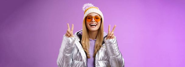 Доброзичлива оптимістична красива блондинка в сонцезахисних окулярах срібна стильна куртка зимовий капелюх показує жест перемоги у мирі, насолоджуючись засніженими канікулами, виходять за межі готелю насолоджуватися лижами, фіолетовим фоном
. - Фото, зображення