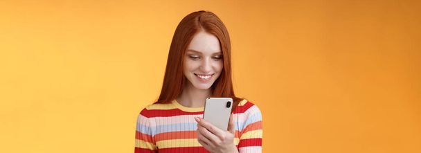 Чарівна сучасна руда дівчина студентка коледжу перевіряє коробку повідомлень, що тримає смартфон, виглядає щасливим усміхненим захопленим дисплеєм мобільних телефонів, отримує сотні подобається фото пост онлайн, помаранчевий фон
. - Фото, зображення